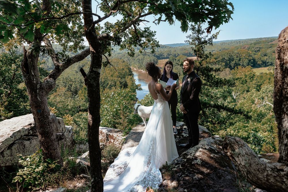 Outdoor wedding ceremony on rocky overlook with bride in white gown, groom, and officiant surrounded by lush forest views.