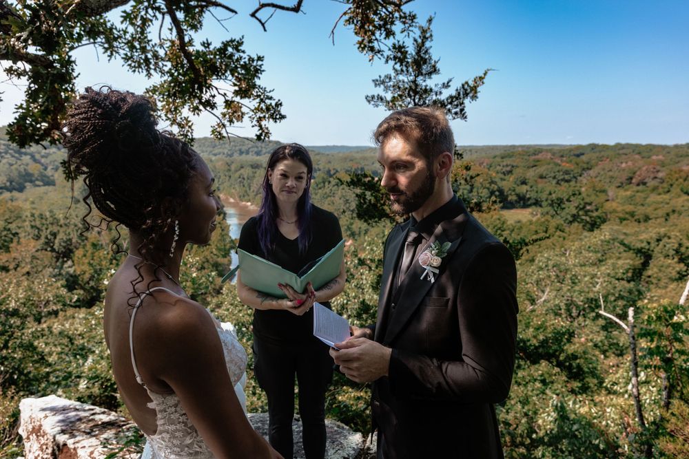 Interracial couple exchanges vows with officiant at scenic overlook wedding ceremony surrounded by lush forest.
