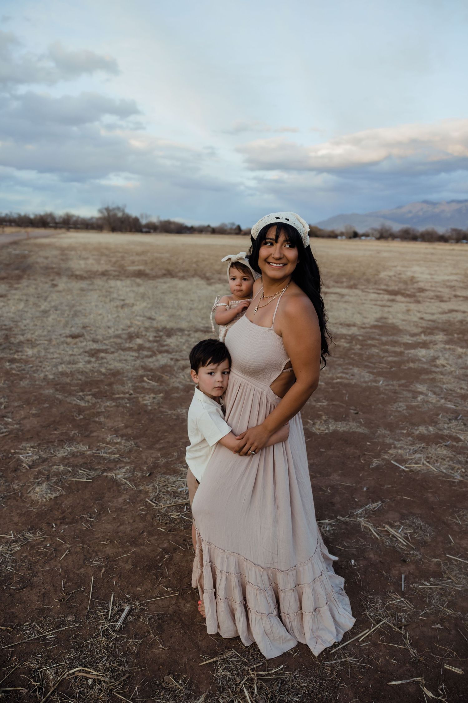A mother in a beige dress stands outdoors with two young children embracing her under a blue sky.