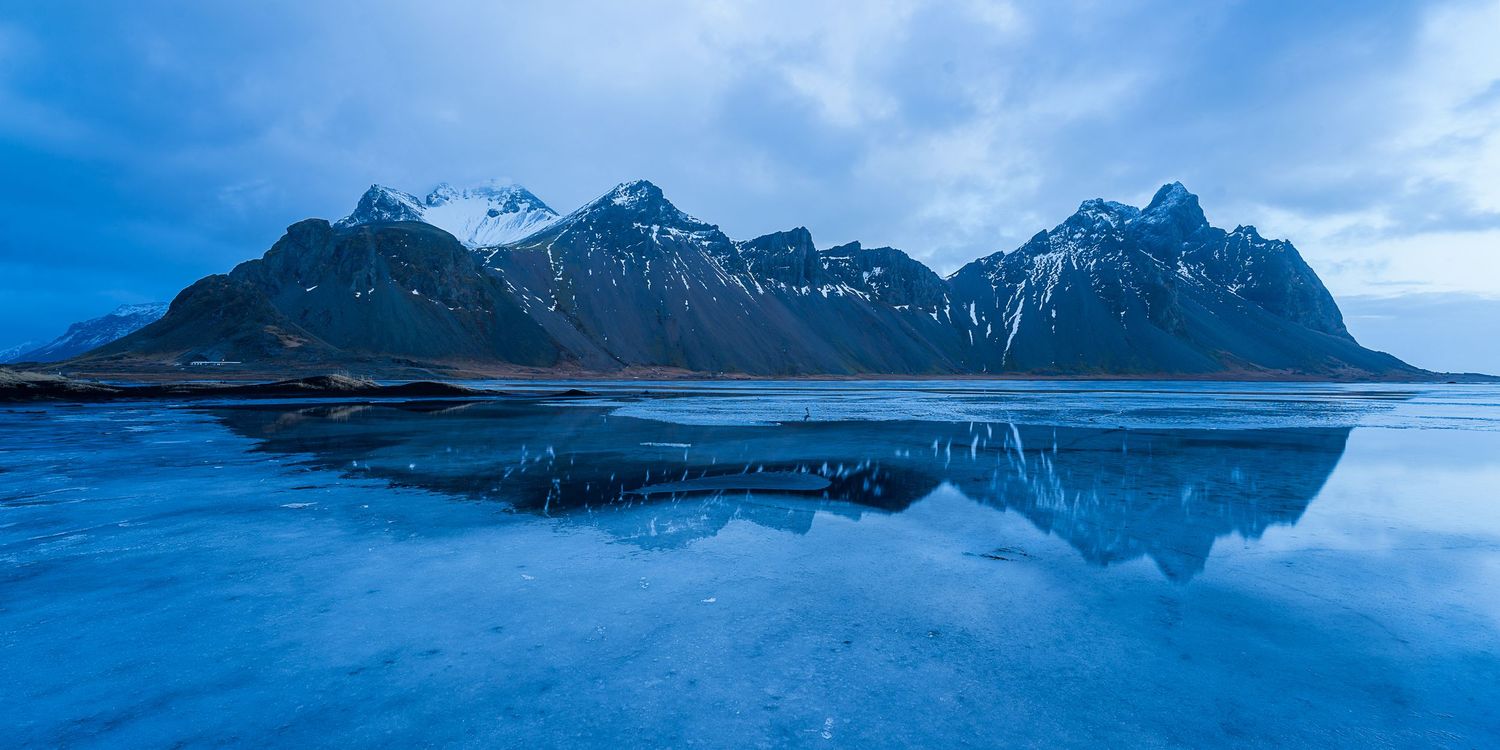 Snow-capped mountains reflect in icy blue waters at dusk in Iceland's dramatic Vestrahorn landscape.