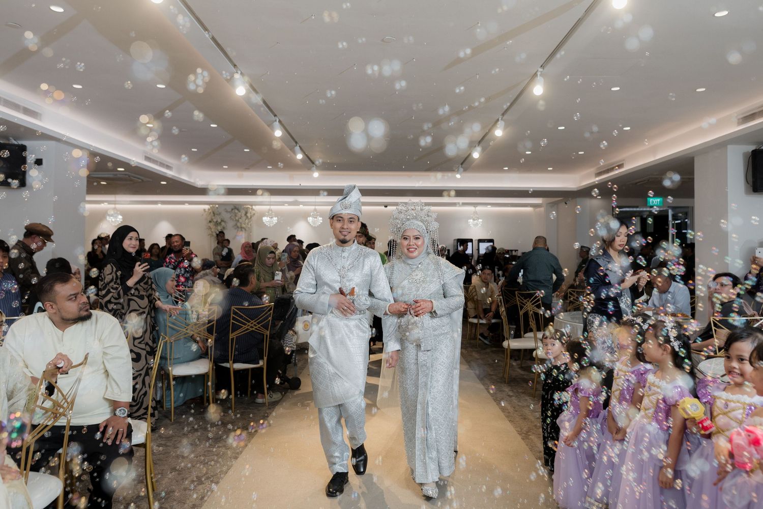 Malay couple in traditional white baju menikah attire posing at elegant wedding reception with floral arch backdrop.