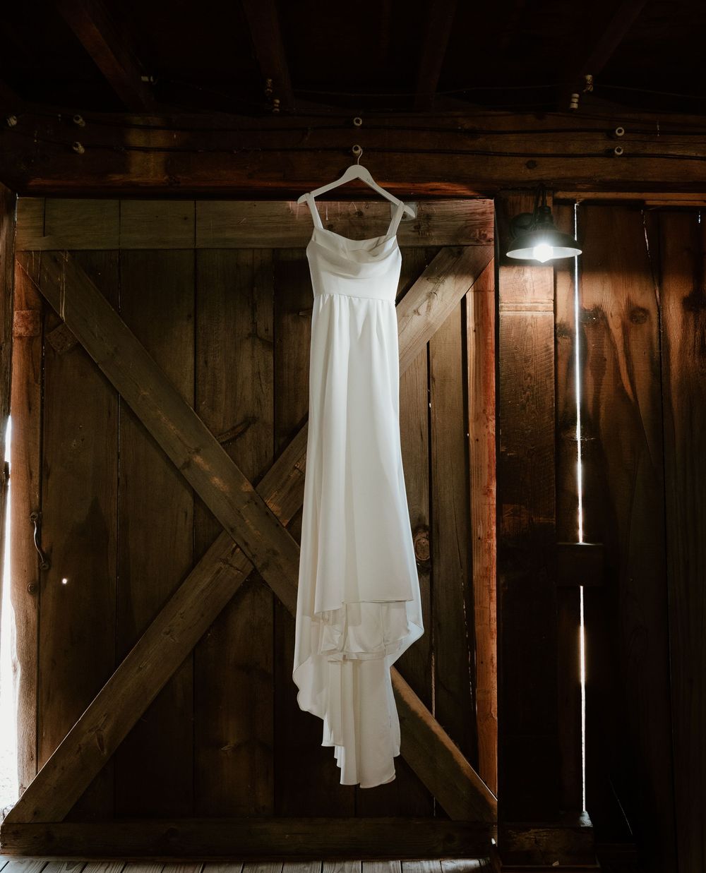 Elegant white wedding dress hanging on a rustic barn door in warm, moody lighting.