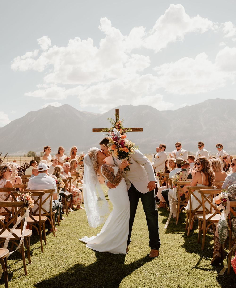 Bride and groom share first kiss at outdoor mountain wedding ceremony with floral cross altar and seated guests.