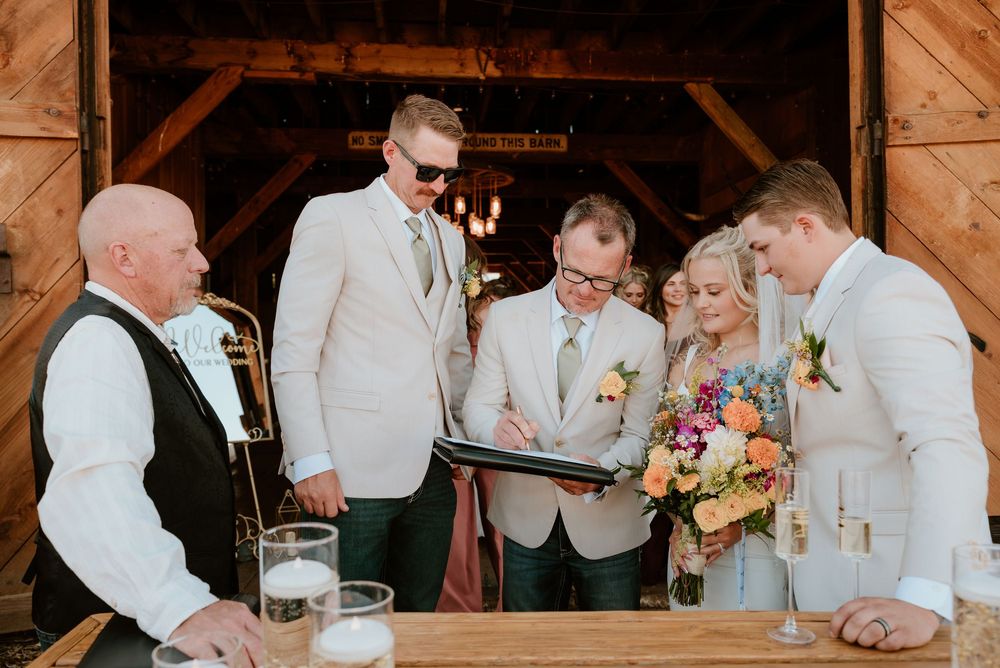 Wedding party signing marriage certificate in rustic barn venue with colorful bridal bouquet.