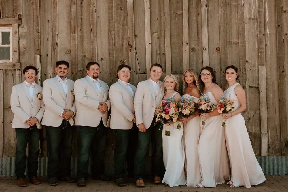 Wedding party poses before rustic barn, groomsmen in tan suits with bridesmaids in blush gowns holding colorful bouquets.