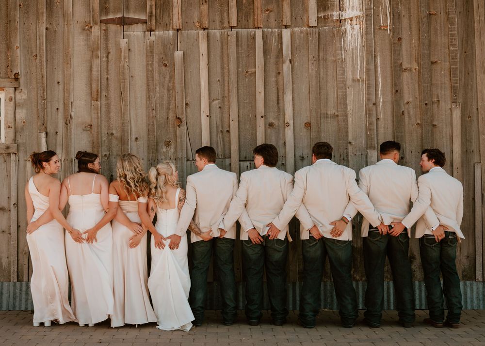 Wedding party in white and dark attire standing together facing a rustic wooden barn wall.