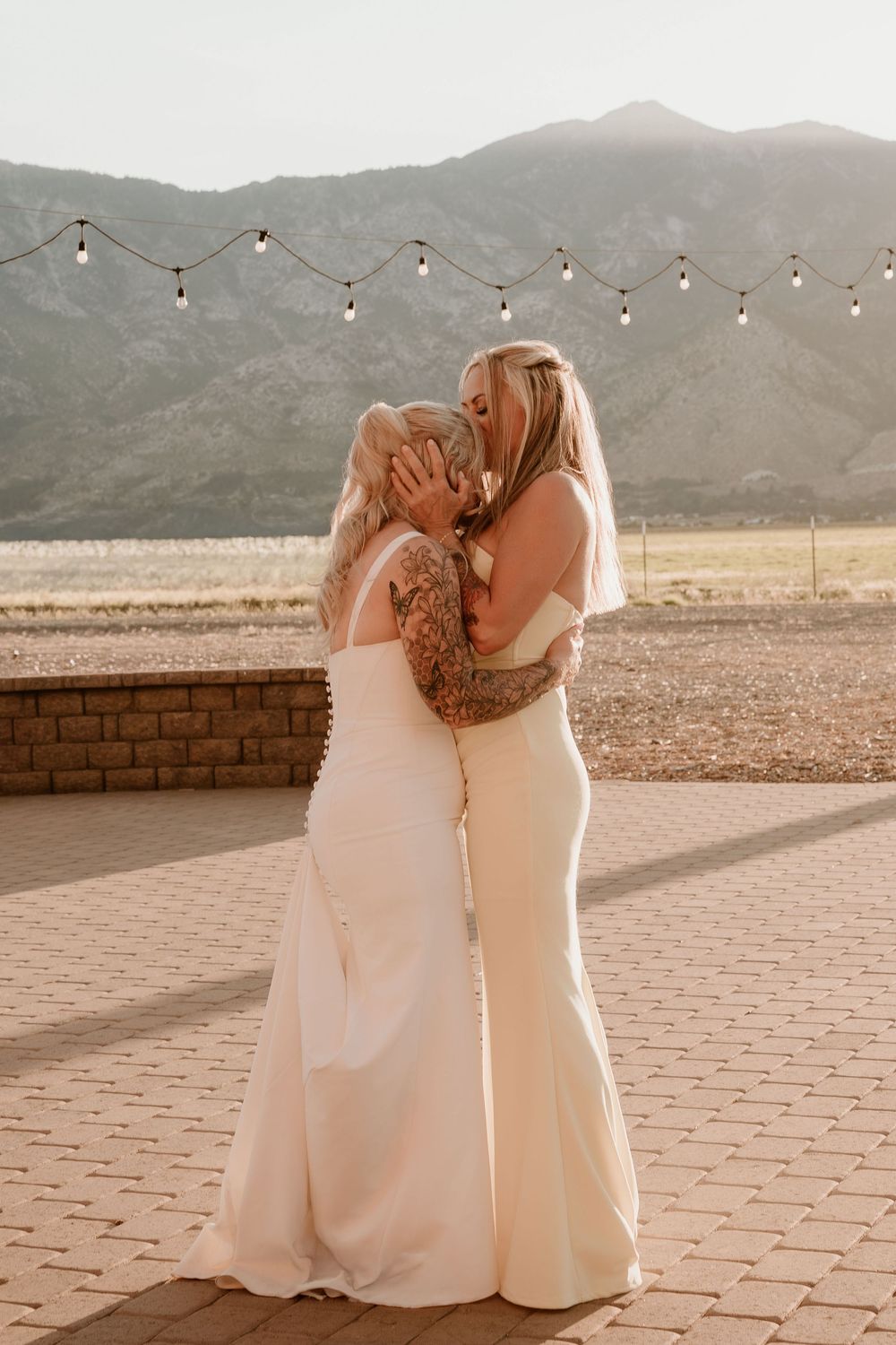 Two brides in white gowns share a kiss at an outdoor wedding with mountain views and string lights.