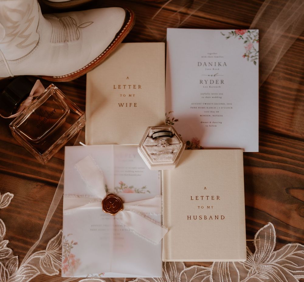Elegant wedding stationery flat lay with vow books, floral invitations, ring box, and perfume on wood.
