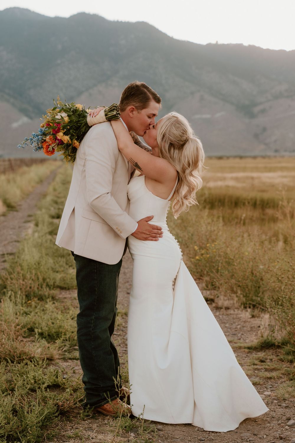 Bride and groom share a romantic kiss in a mountain meadow, she holds a colorful bouquet in her white gown.