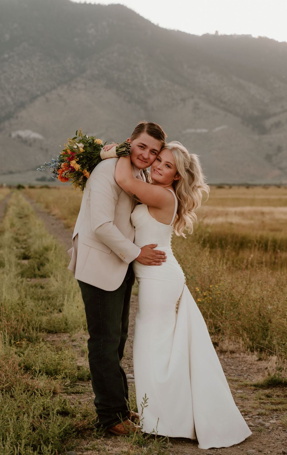 Bride and groom embrace in a field with mountain backdrop, she in white gown, he in tan suit holding colorful bouquet.