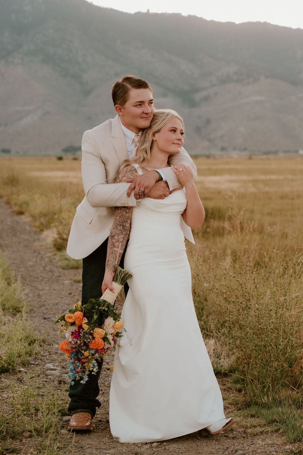A bride and groom embrace on a dirt path with golden fields and mountains in the background during their outdoor wedding.