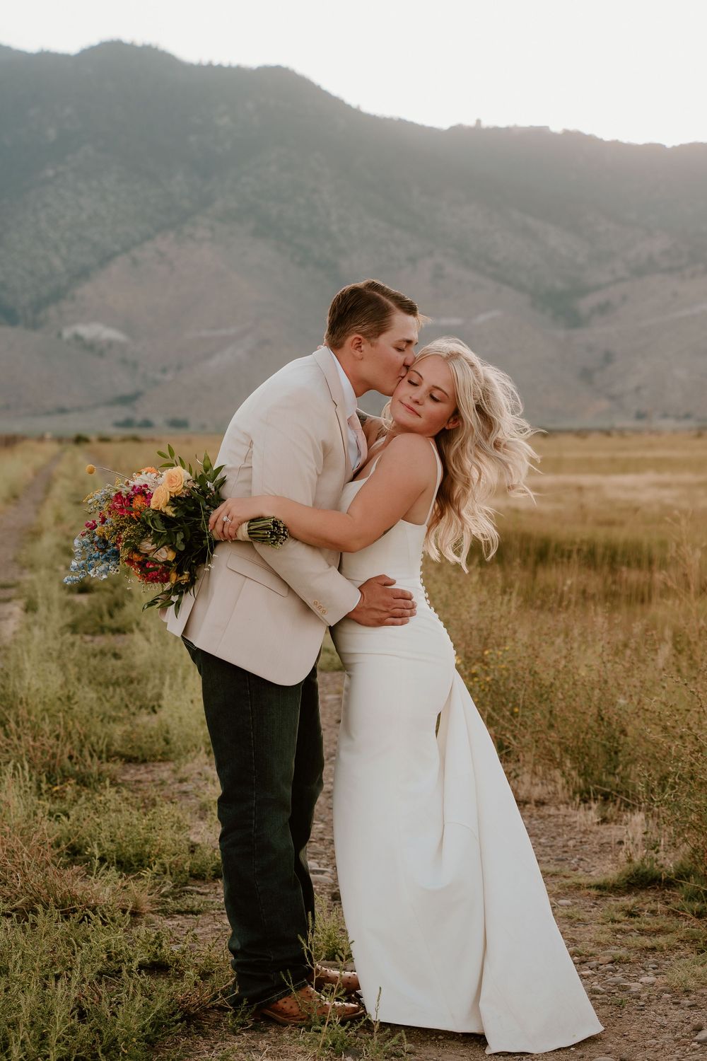 Groom in beige suit kisses bride in white gown holding colorful bouquet with mountain backdrop at golden hour.