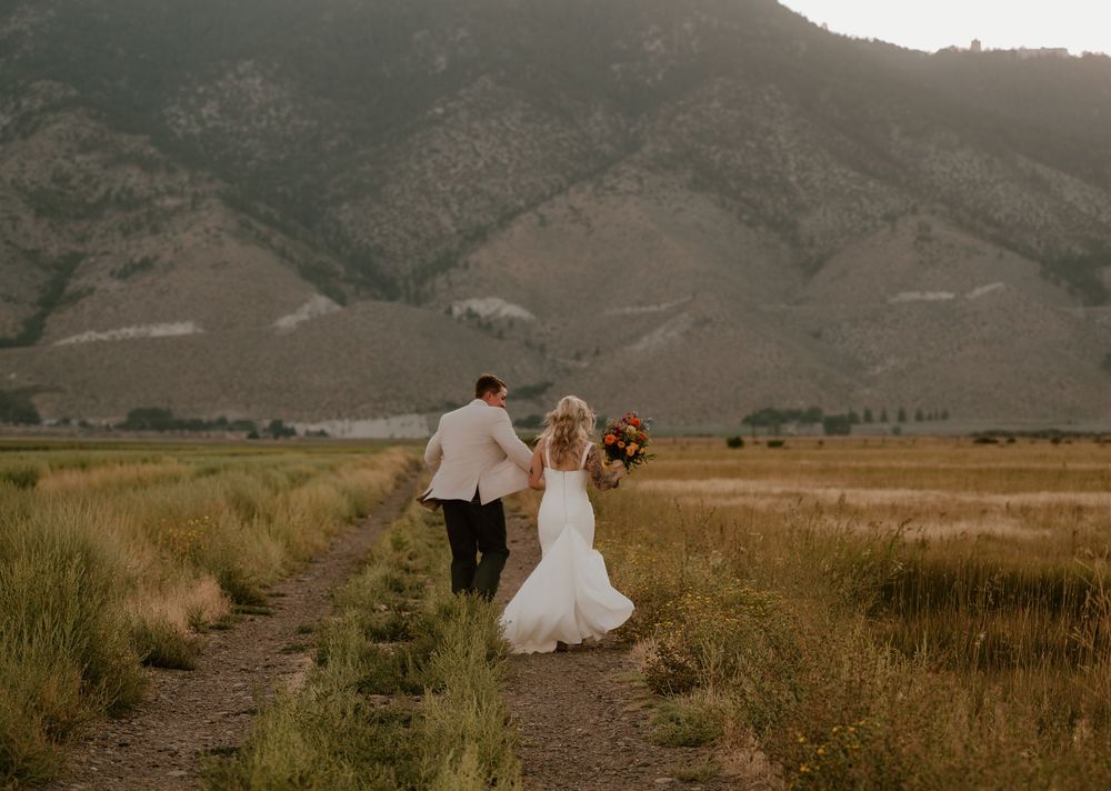 A bride and groom walk hand-in-hand through a mountain meadow path on their wedding day.