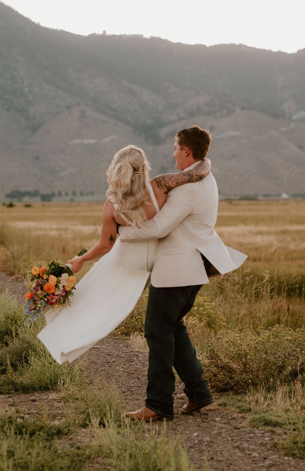 Groom lifting bride in white dress with colorful bouquet, mountains and golden fields in background.