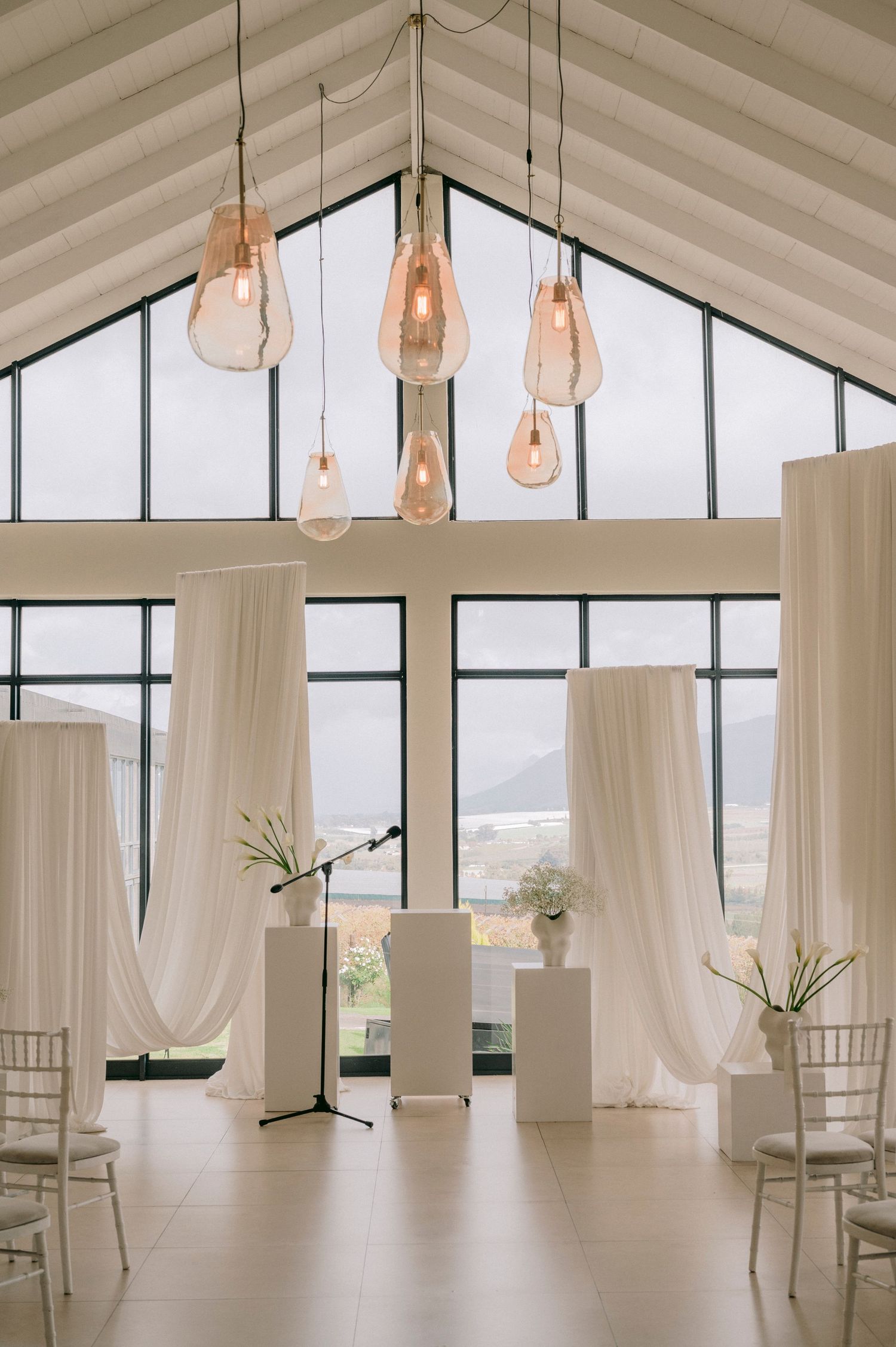 Elegant wedding ceremony venue with white draping, pendant lights, and floor-to-ceiling windows overlooking scenic mountains.