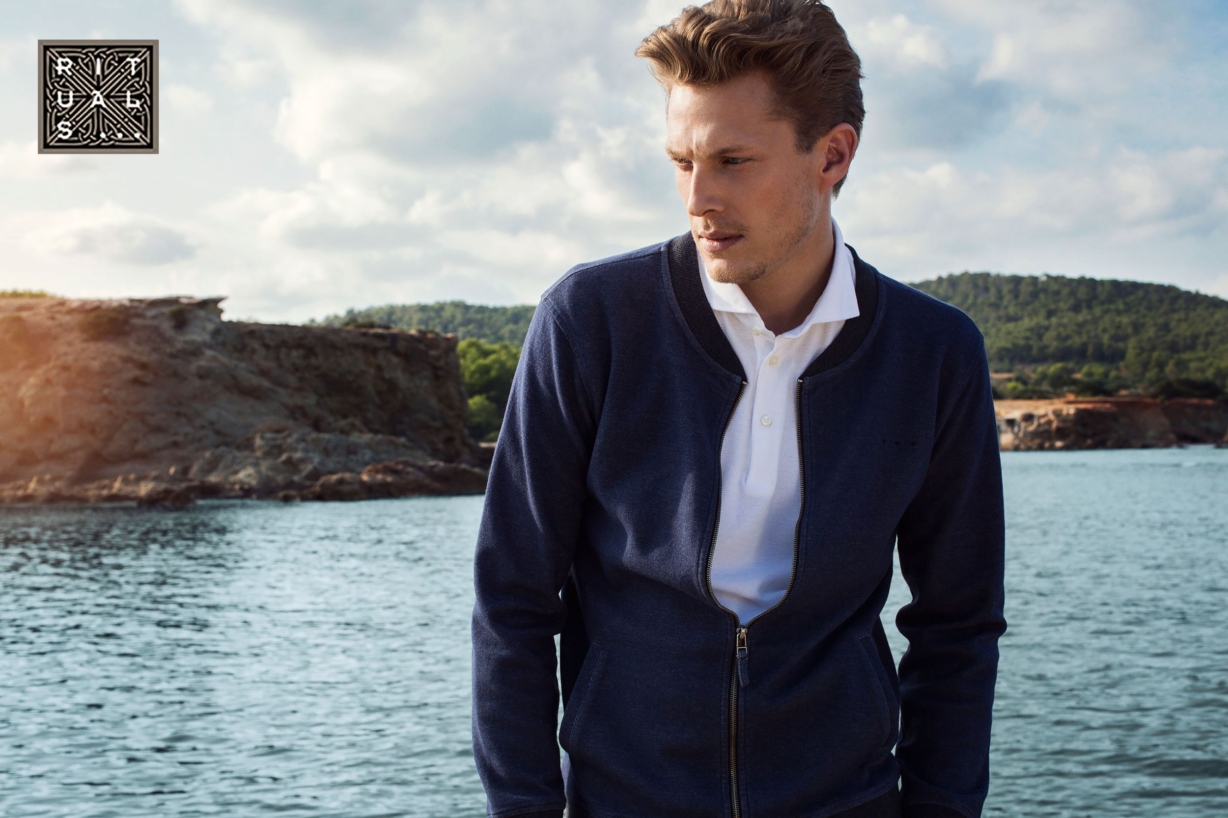 Man in navy zip-up jacket standing near water in outdoor fashion campaign.