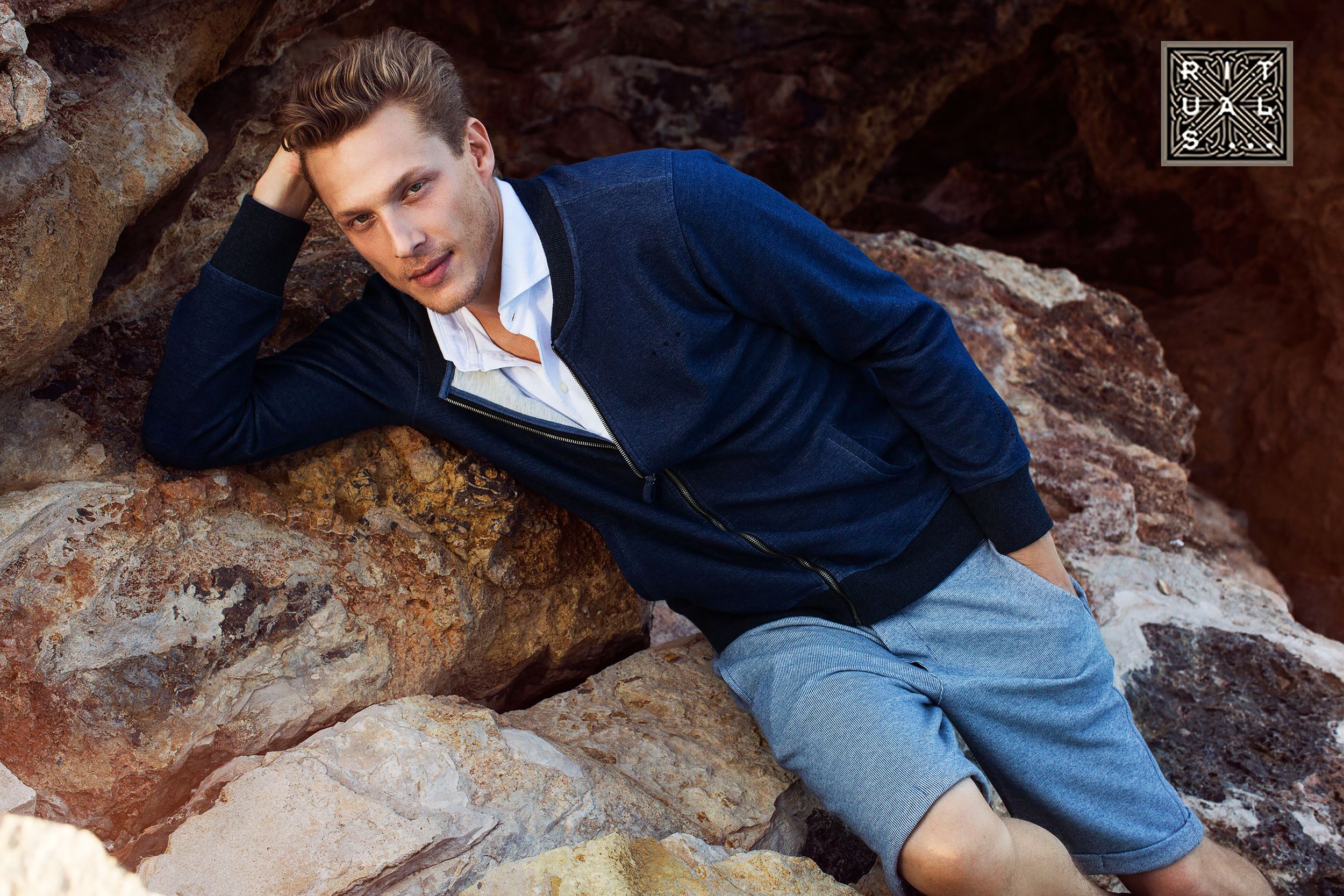 Man in navy cardigan and grey shorts relaxes on rustic red rocks outdoors.