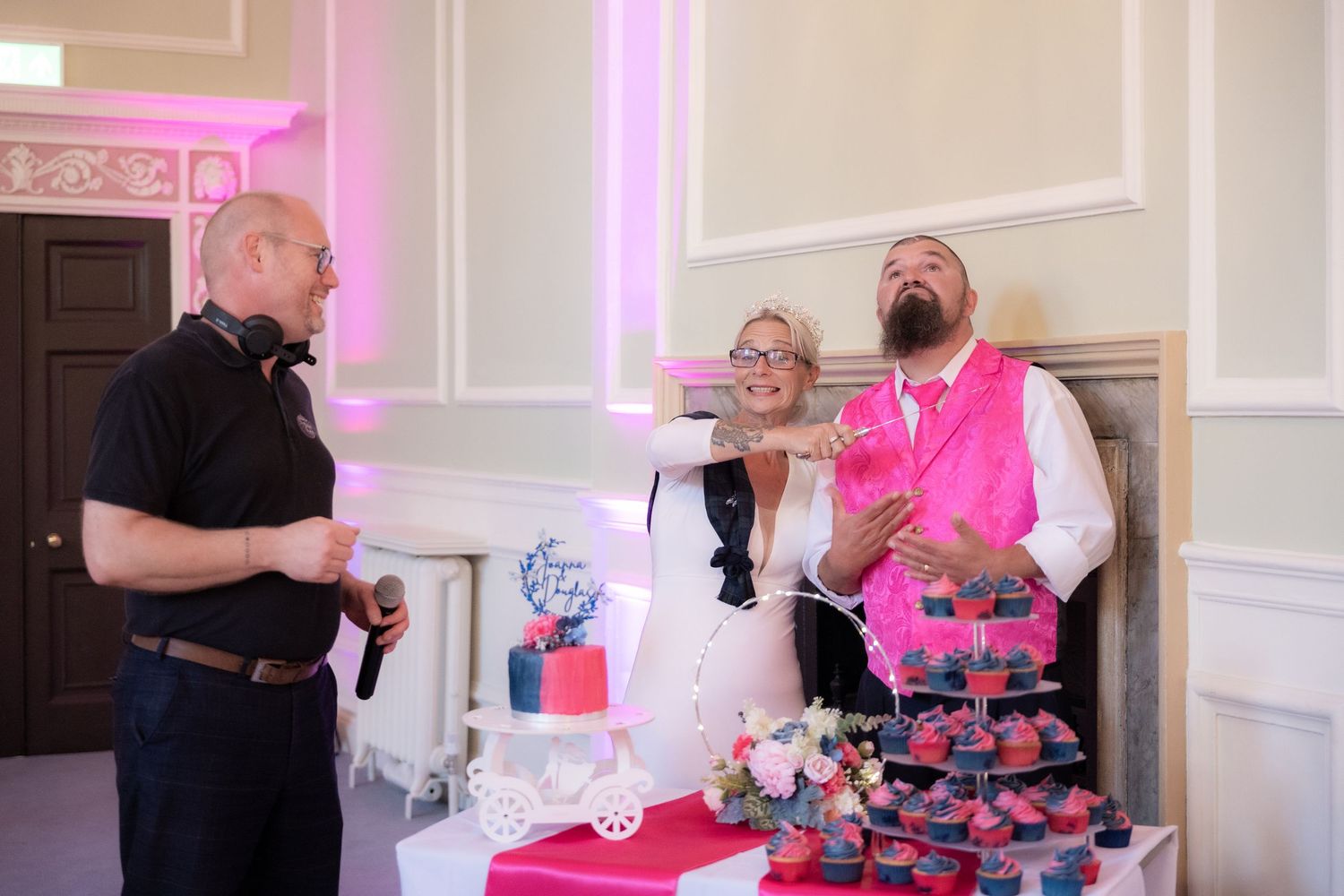 Bride and groom laughing with guest near elegant wedding cake display with pink cupcakes at reception.
