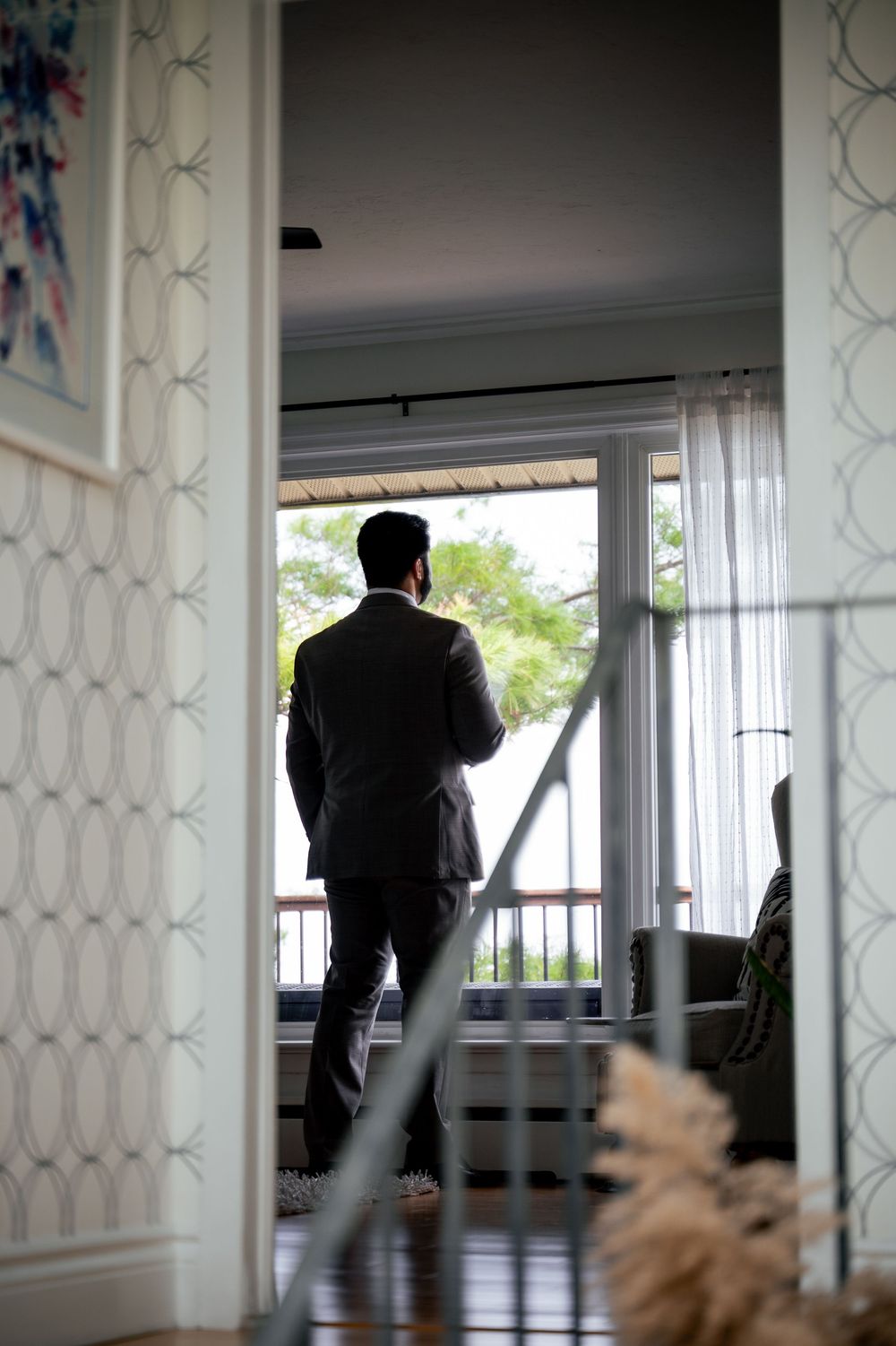 Man in suit standing by large window with sheer curtains, viewed from hallway in elegant home interior.