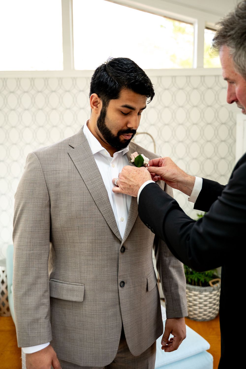 An older man pins a boutonniere on a bearded groom in a tan suit before a wedding ceremony.