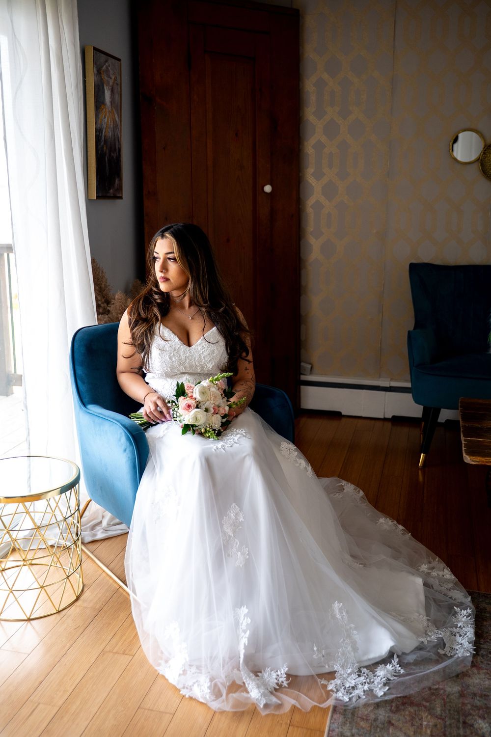 Bride in white lace wedding gown holding floral bouquet seated in blue velvet chair by window.