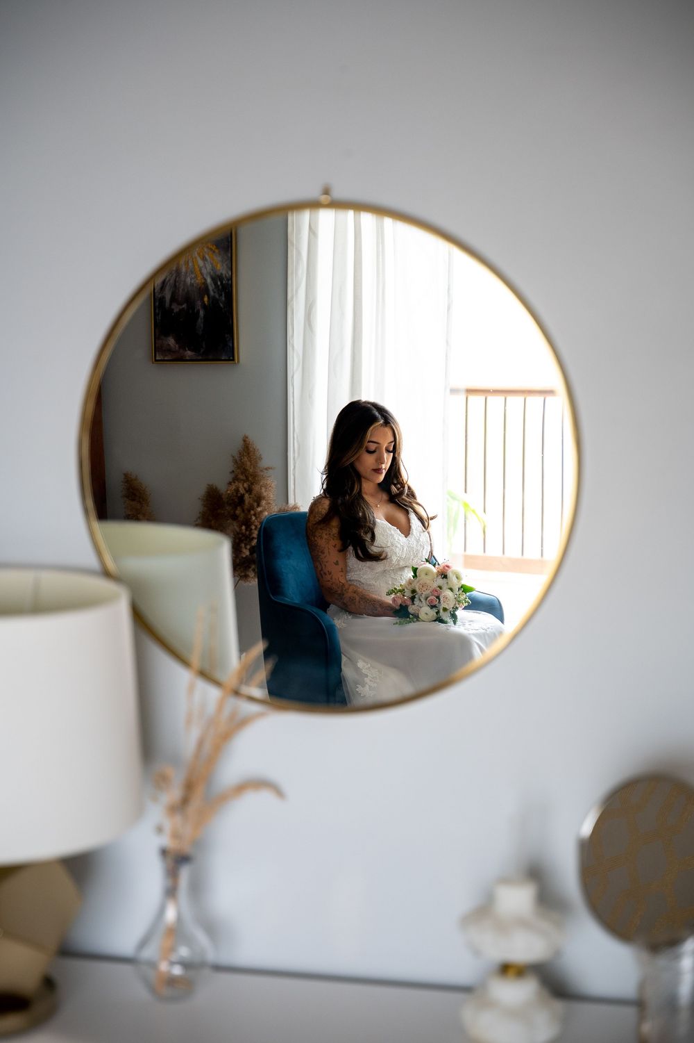 Bride in white wedding dress holding bouquet reflected in round gold-framed mirror on white wall.