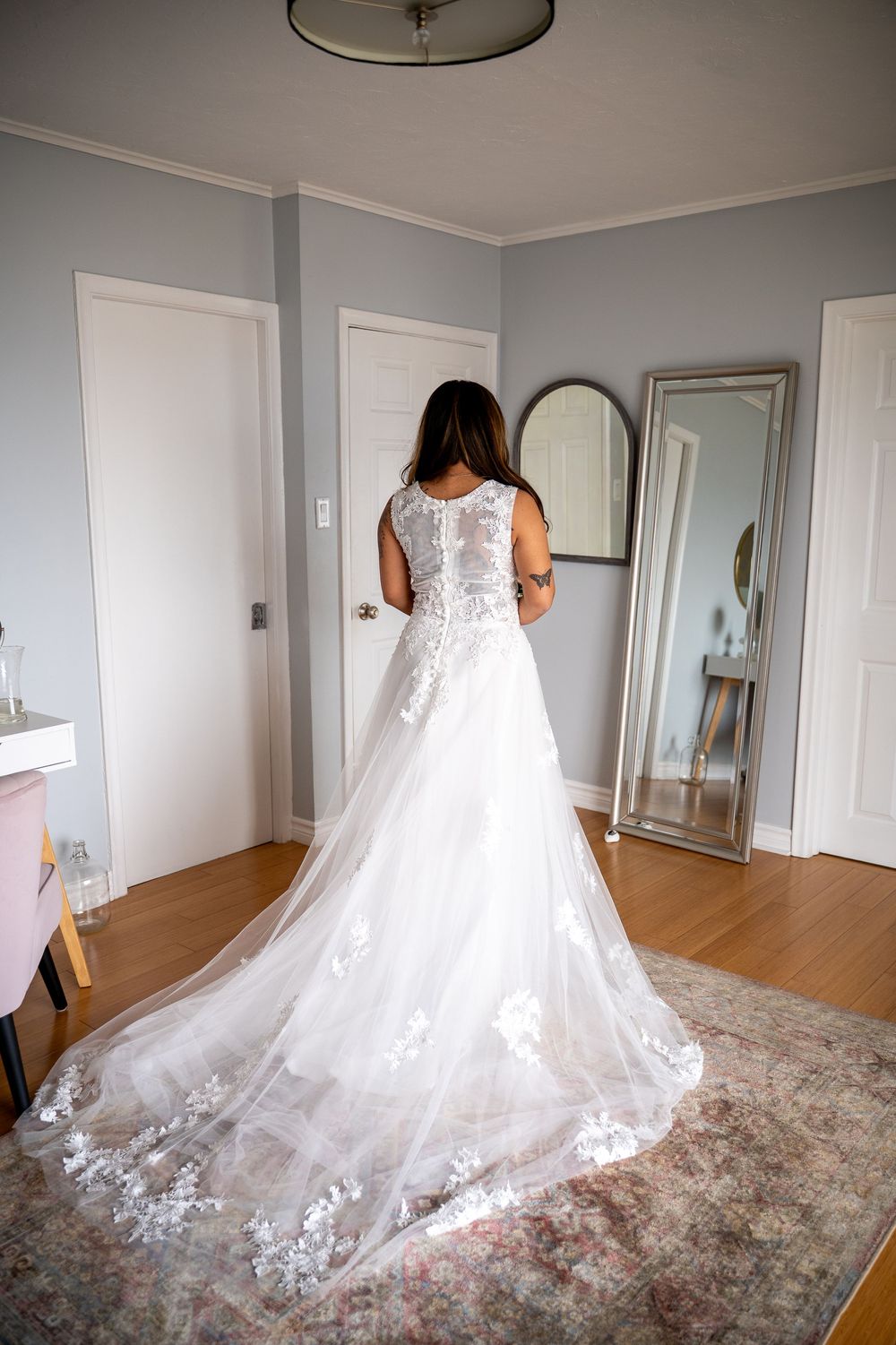 Bride in white lace A-line wedding gown with long train viewed from behind standing in bedroom.