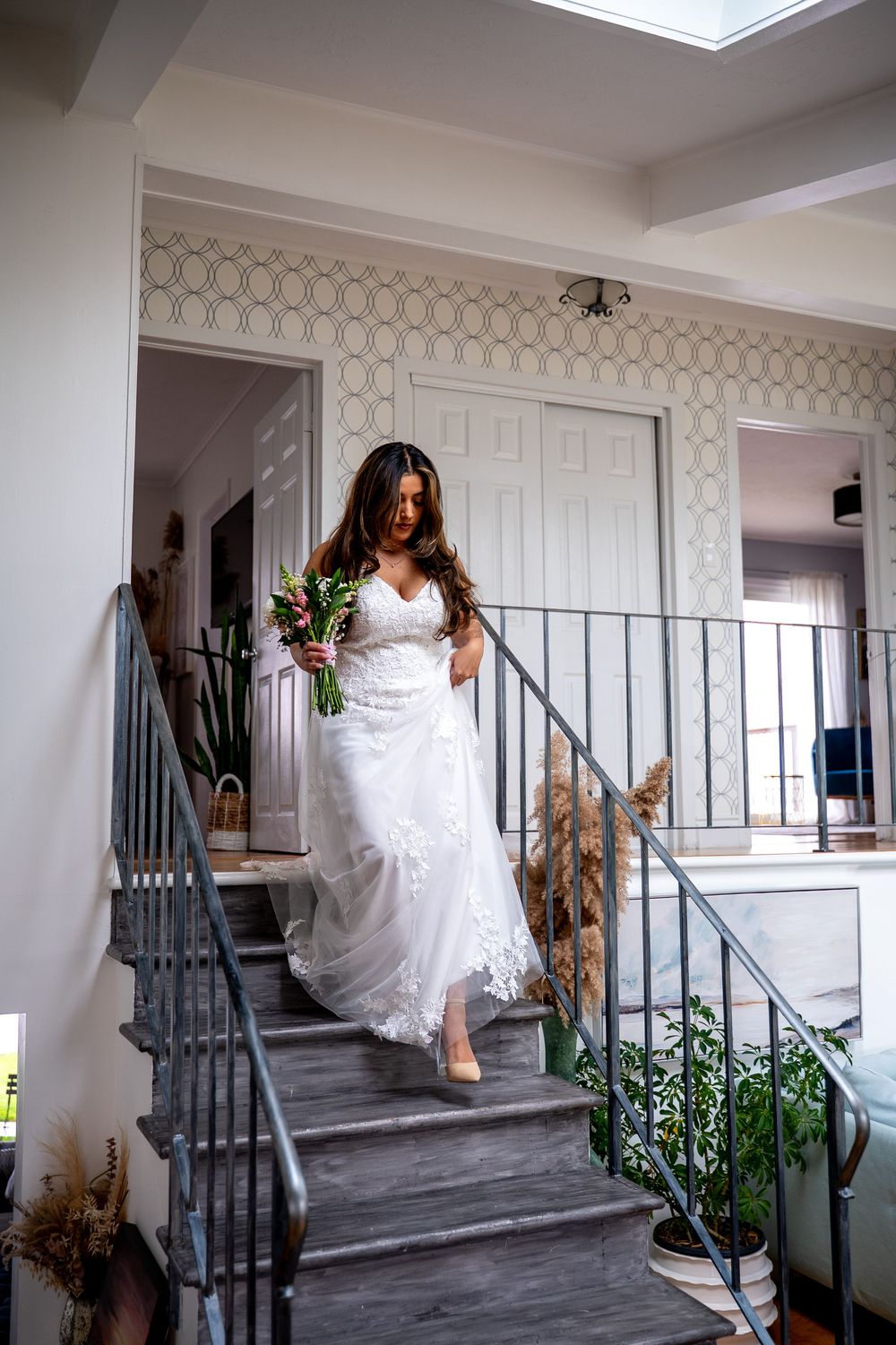 Bride in white lace gown descending staircase holding bouquet in elegant home interior.