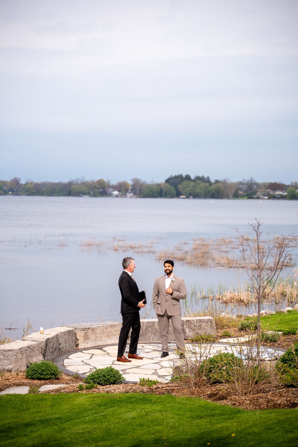 Two men in suits exchange vows outdoors on a stone patio overlooking a serene lake at a wedding ceremony.