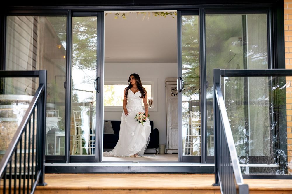 Bride in white wedding dress holding bouquet stands in open glass doors of modern home exterior.