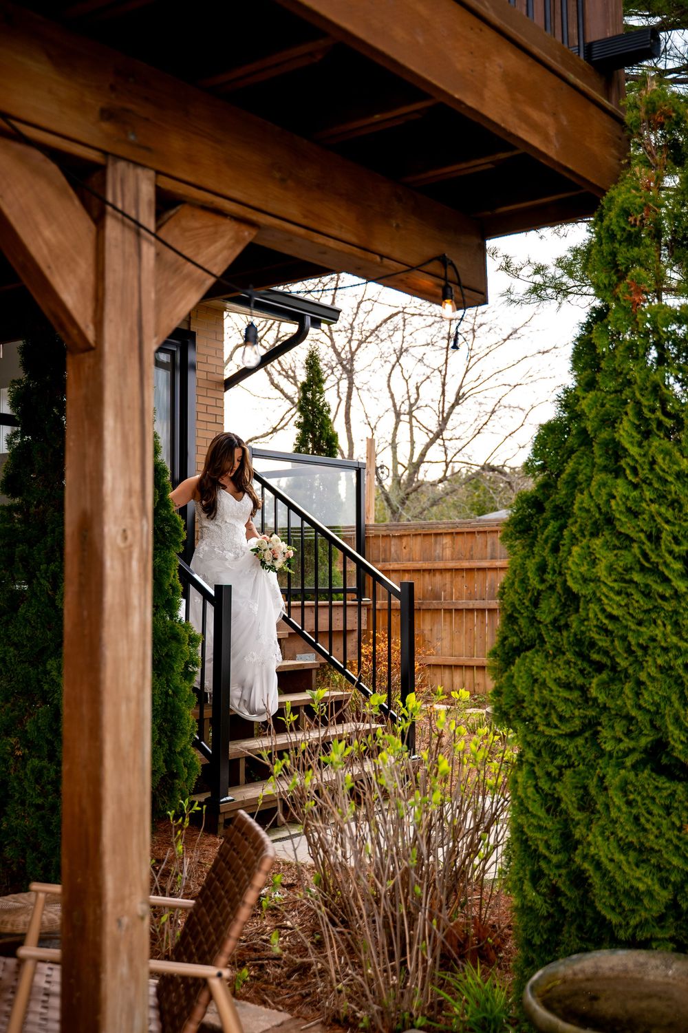 Bride in white lace gown descending outdoor staircase surrounded by evergreen trees at wedding venue.
