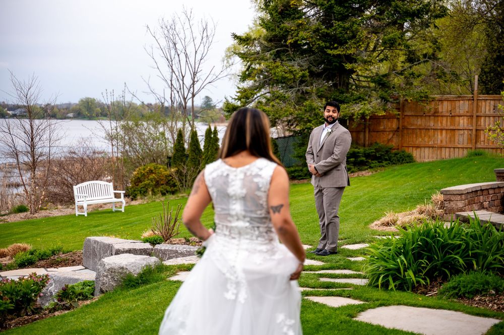 Bride in white lace gown walks toward groom in gray suit during outdoor first look ceremony by waterfront garden.