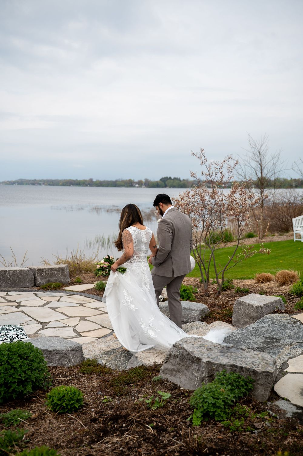 Bride and groom walking toward lakeside garden on stone path on cloudy wedding day.