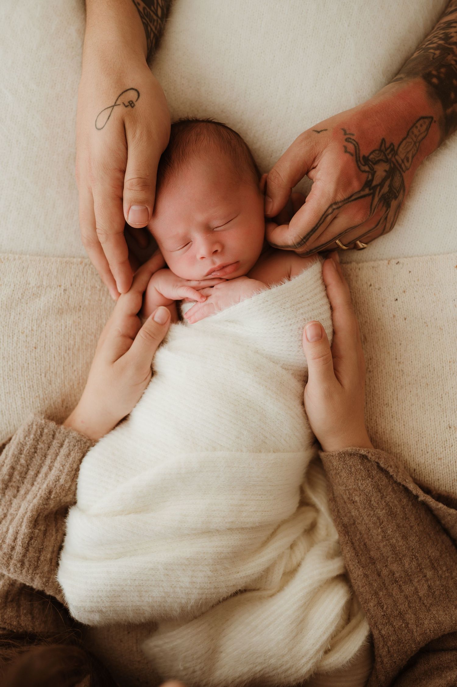 Sleeping newborn baby in cream knit outfit cradled gently by parents hands in a serene newborn photography portrait.