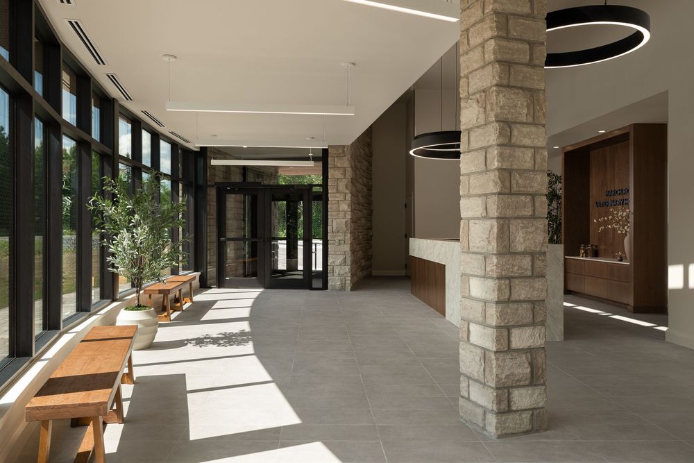Modern building lobby with stone columns, floor-to-ceiling windows, wooden benches, and circular pendant lights on polished floors.