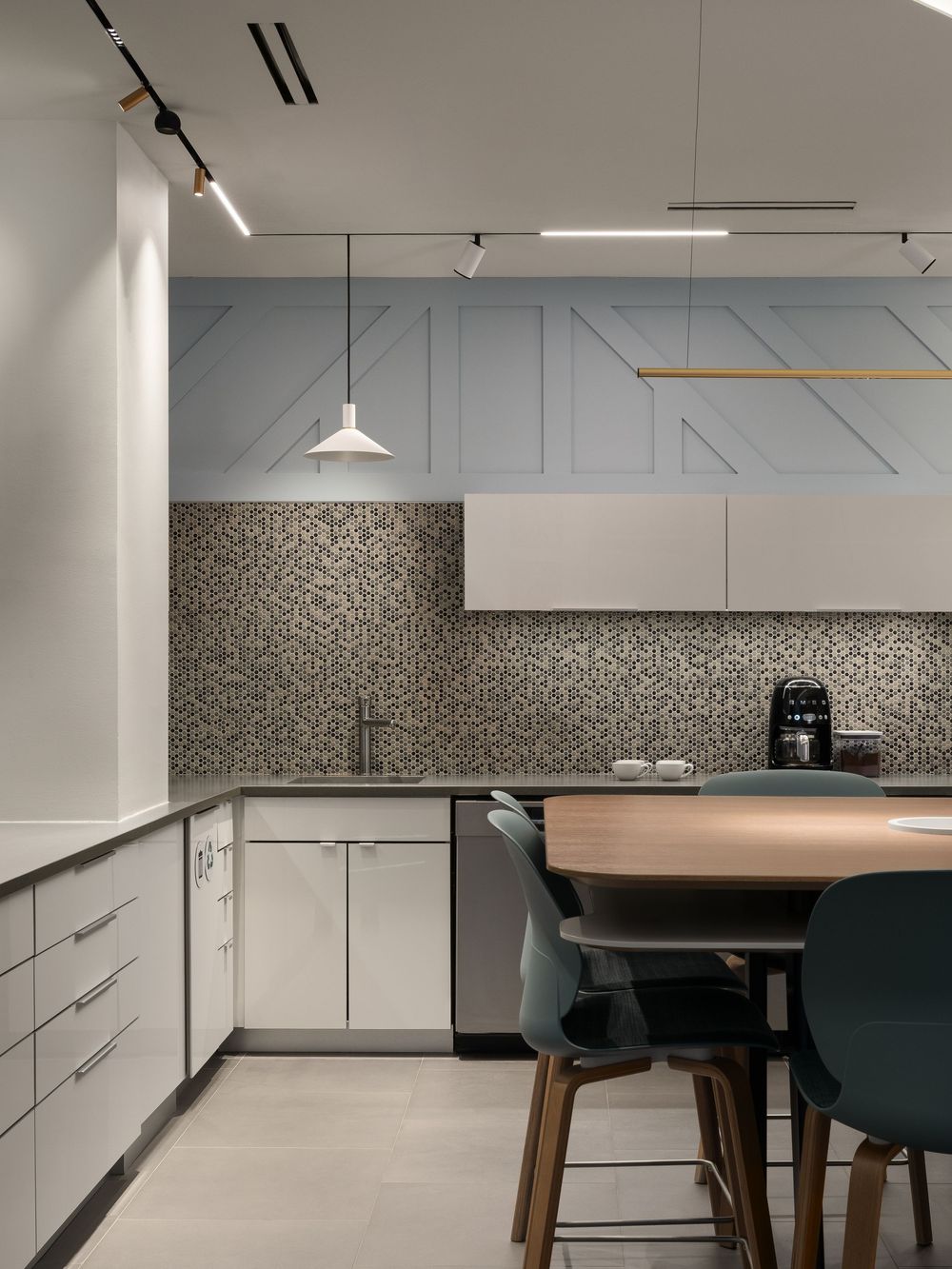 Modern kitchen with mosaic tile backsplash, white cabinets, round wood dining table, teal chairs, and sleek pendant lighting.
