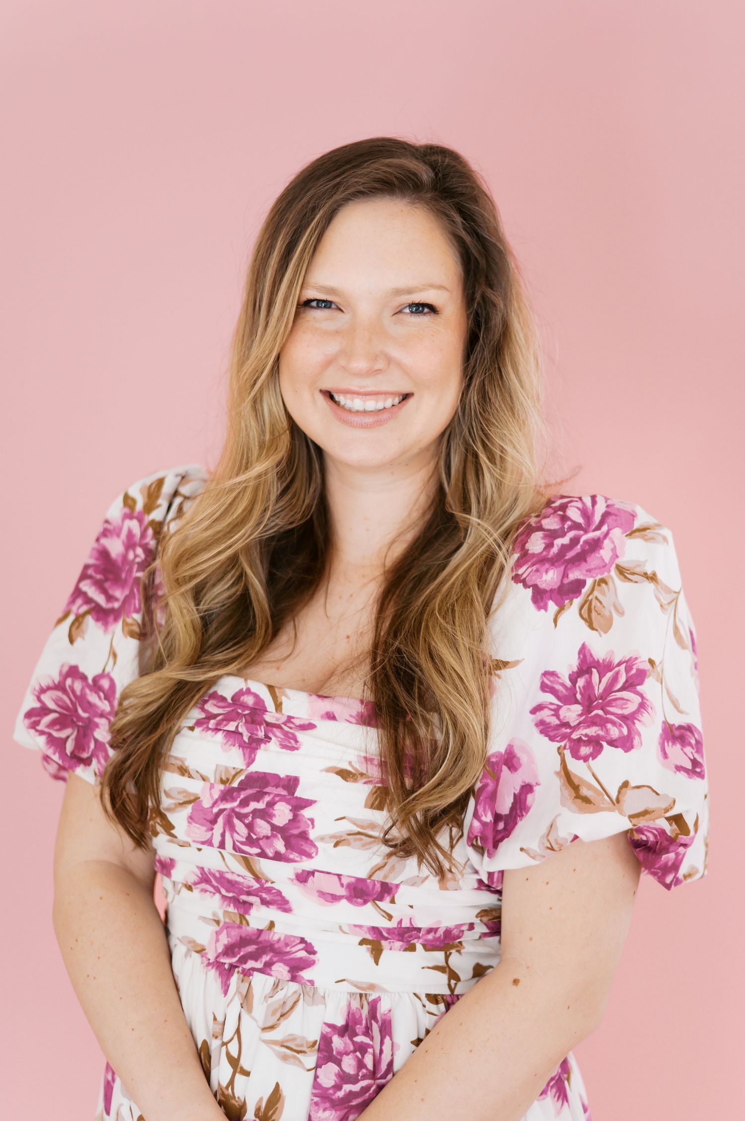 Smiling woman with long blonde hair wearing a white floral dress with pink flowers against a pink background.