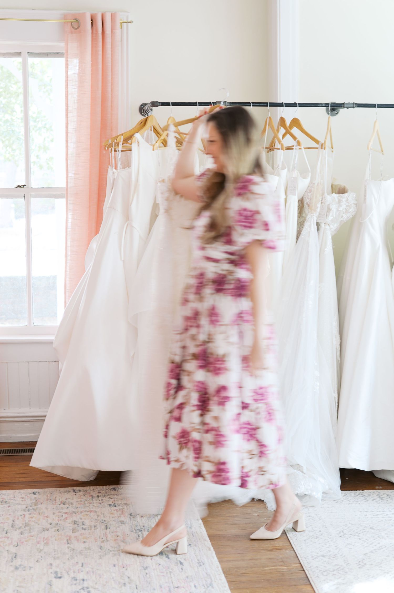 Woman in floral dress browsing white wedding gowns on a rack in a bright bridal boutique.