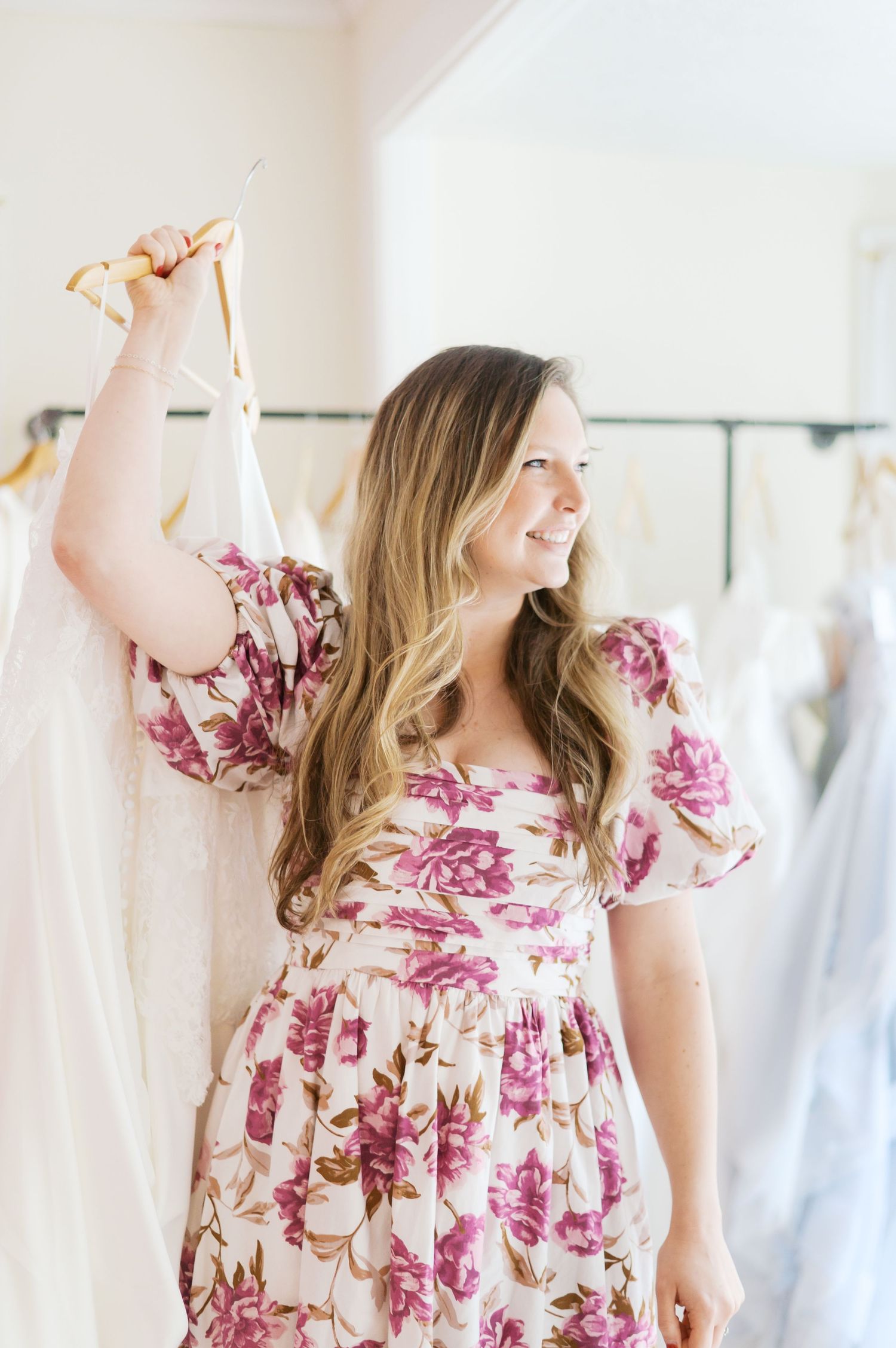 Woman in floral dress holding wedding gown hanger in bridal boutique, smiling among white dresses.
