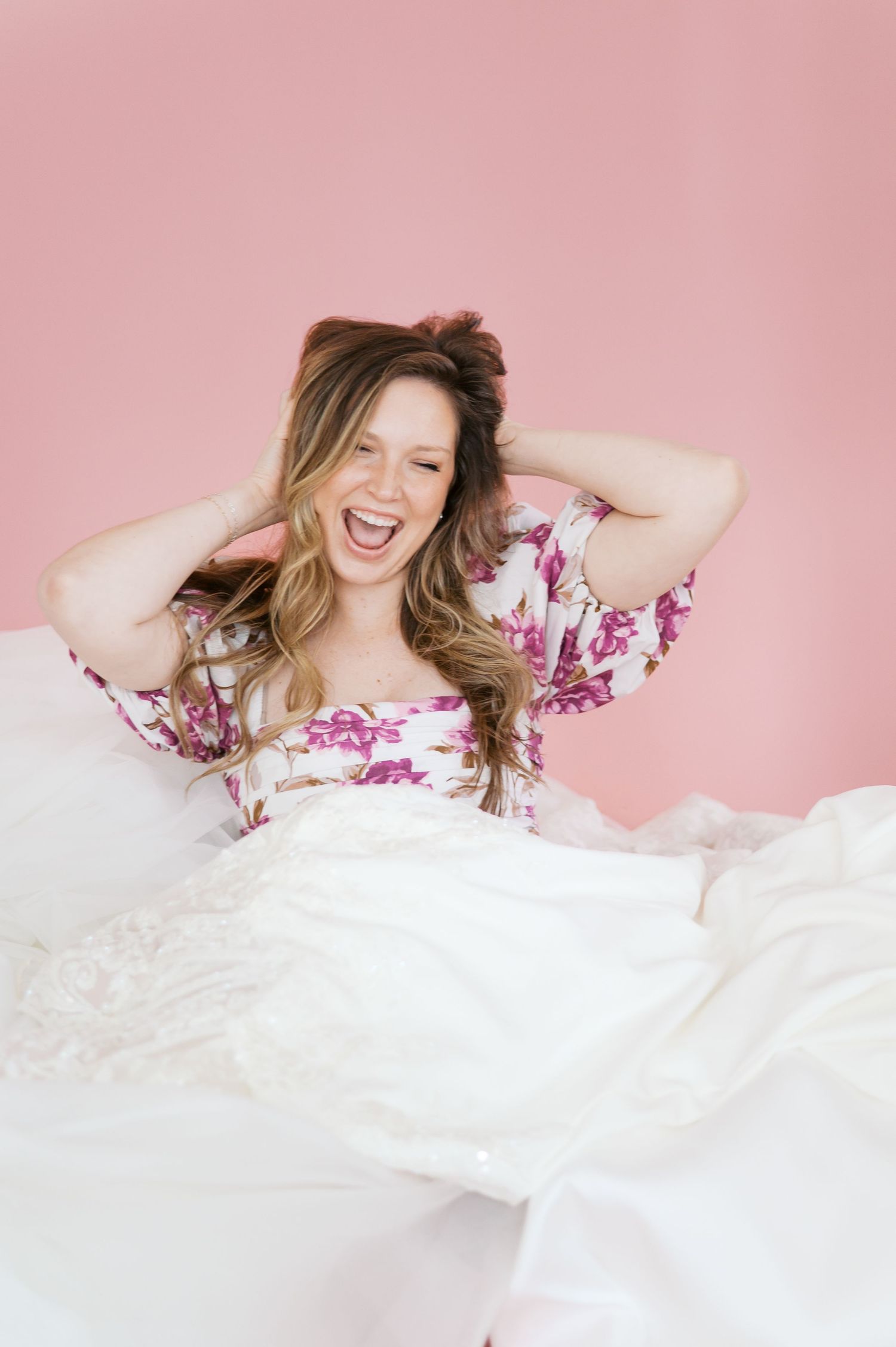Laughing bride in floral robe surrounded by white wedding dress skirt against pink background.