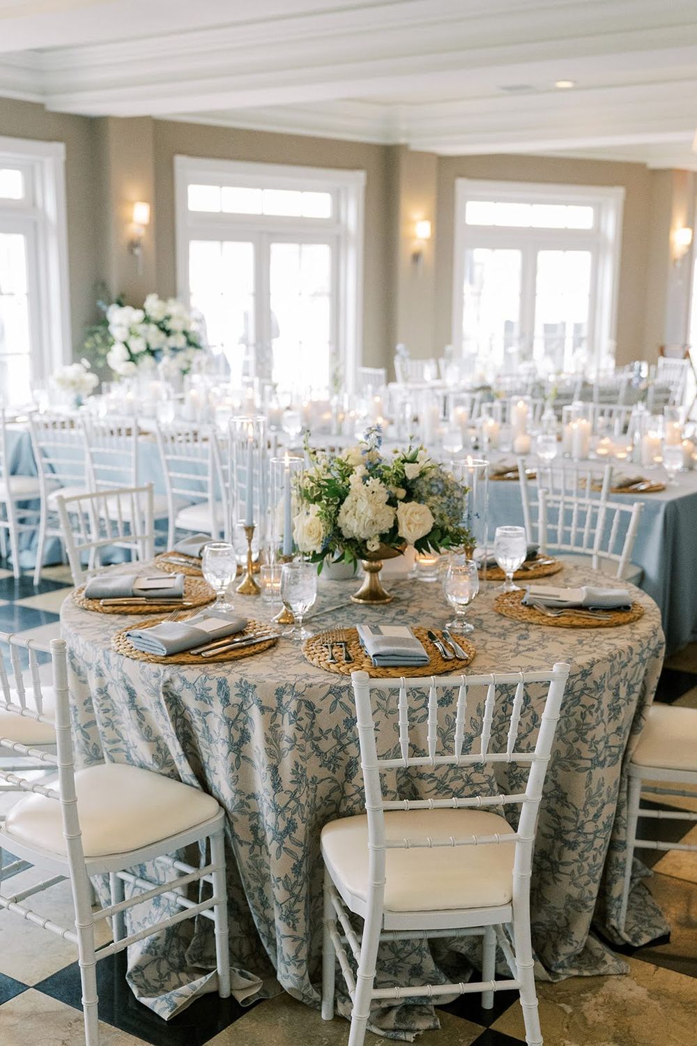 Elegant wedding reception with blue floral linens, gold chargers, white floral centerpieces, and chiavari chairs in a bright ballroom.