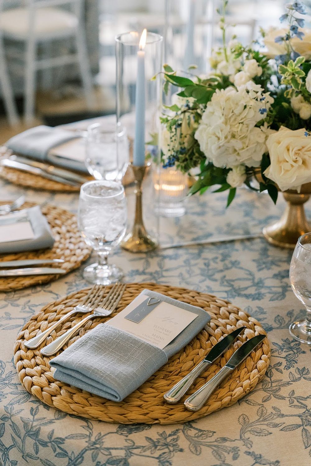 Elegant wedding table setting with woven chargers, blue napkins, crystal glassware, candles, and white floral centerpiece.