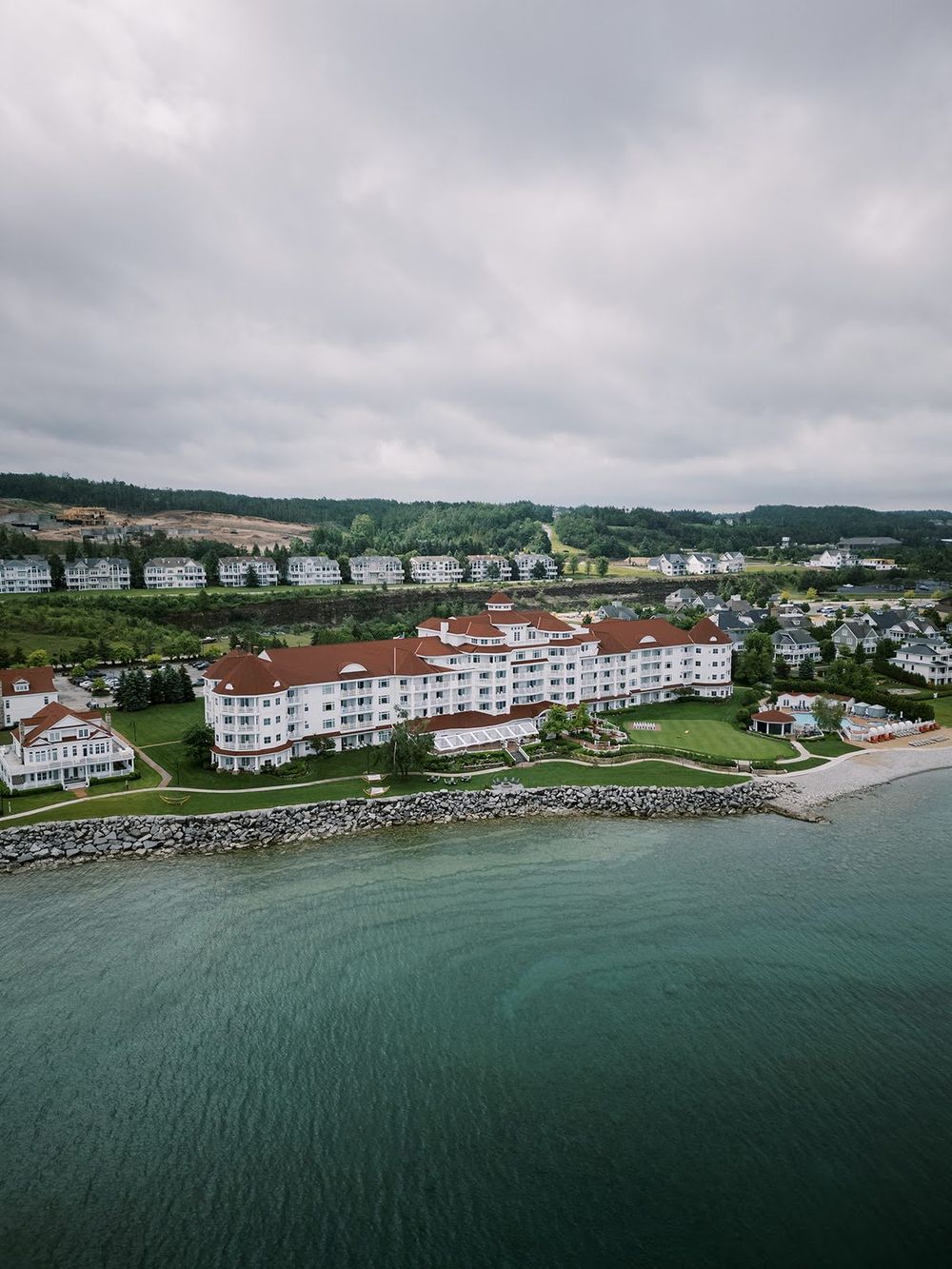Aerial view of a large white waterfront hotel with red roof near rocky shoreline under overcast skies.