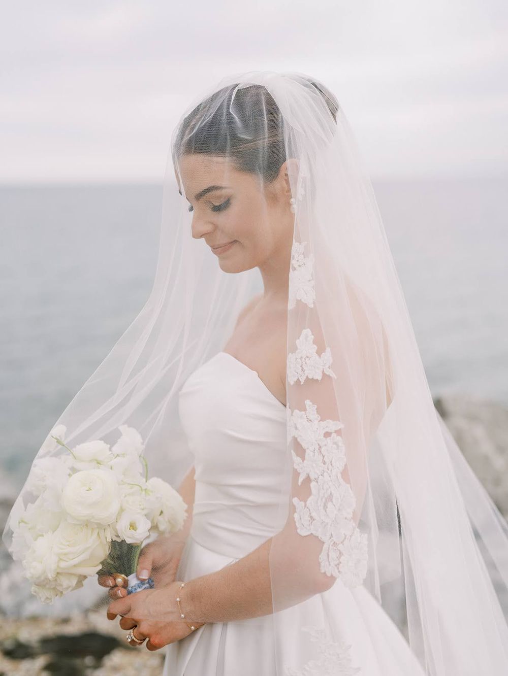 Bride in strapless white gown and lace-trimmed veil holds white bouquet by the ocean.