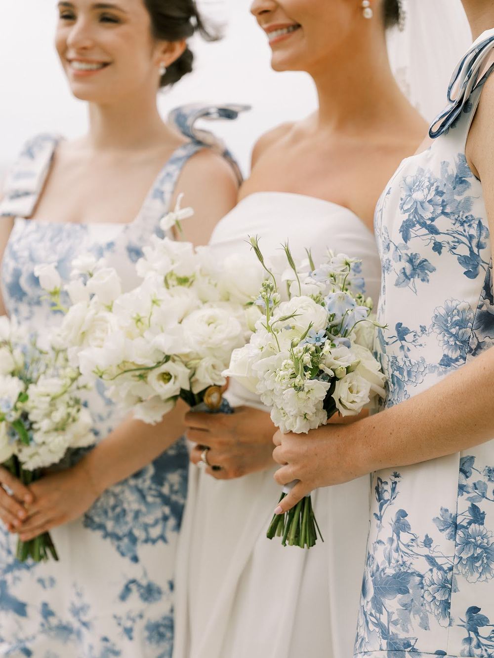 Bride and bridesmaids in blue floral dresses holding white bouquets of ranunculus and roses at a wedding.