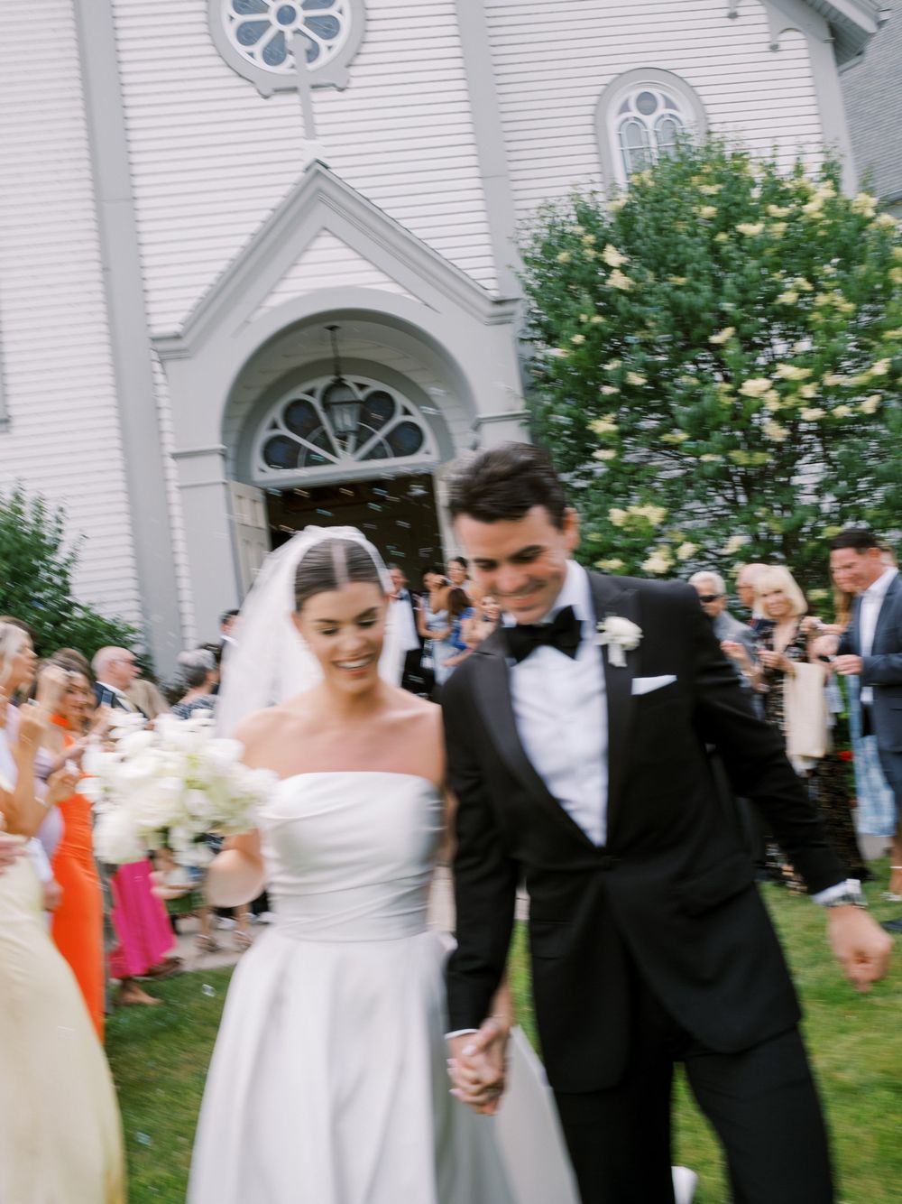 Bride and groom exit white church holding hands while guests celebrate with confetti on sunny wedding day.