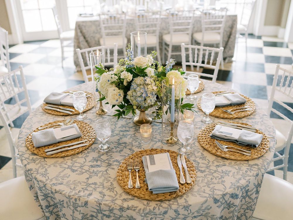 Elegant wedding reception table with gold chargers, blue floral linens, white floral centerpiece, and chiavari chairs.
