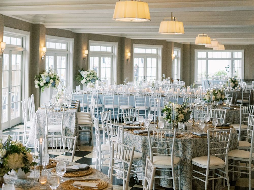 Elegant wedding reception hall with white Chiavari chairs, floral centerpieces, gold-accented tables, and large windows with natural light.