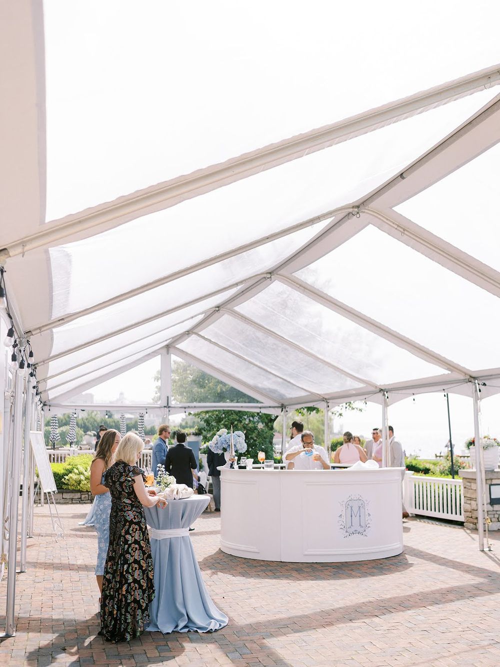 Elegant outdoor wedding reception under a clear tent with guests mingling around a monogrammed bar on a sunny day.