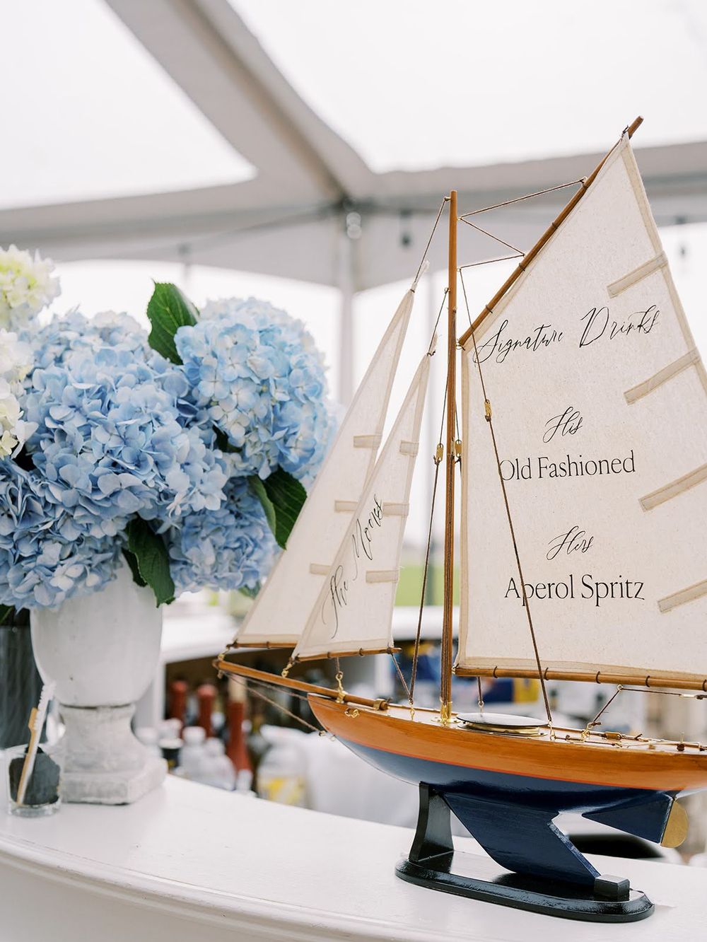 Nautical wedding decor with a model sailboat displaying signature drinks menu beside blue hydrangeas in a white vase.